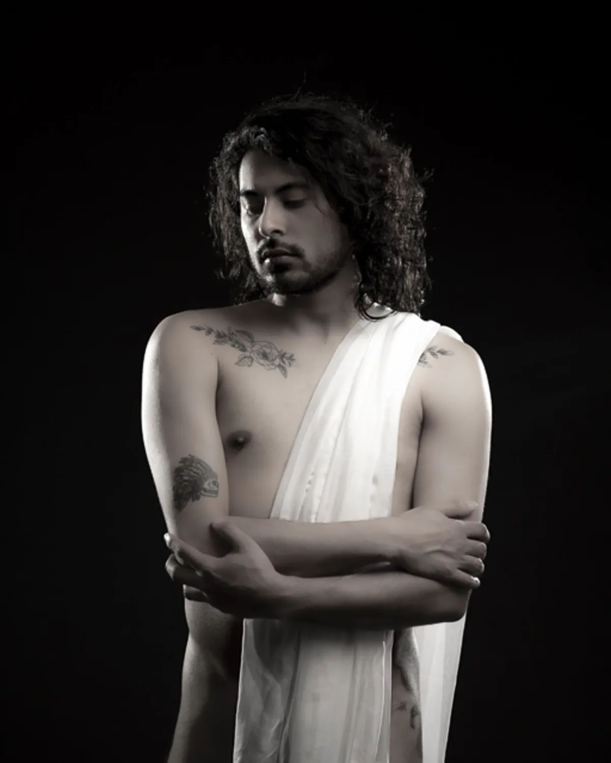 Latino man standing in profile wearing a white silk wrap, gazing downward
with arms crossed over chest in an elegant low-key studio portrait