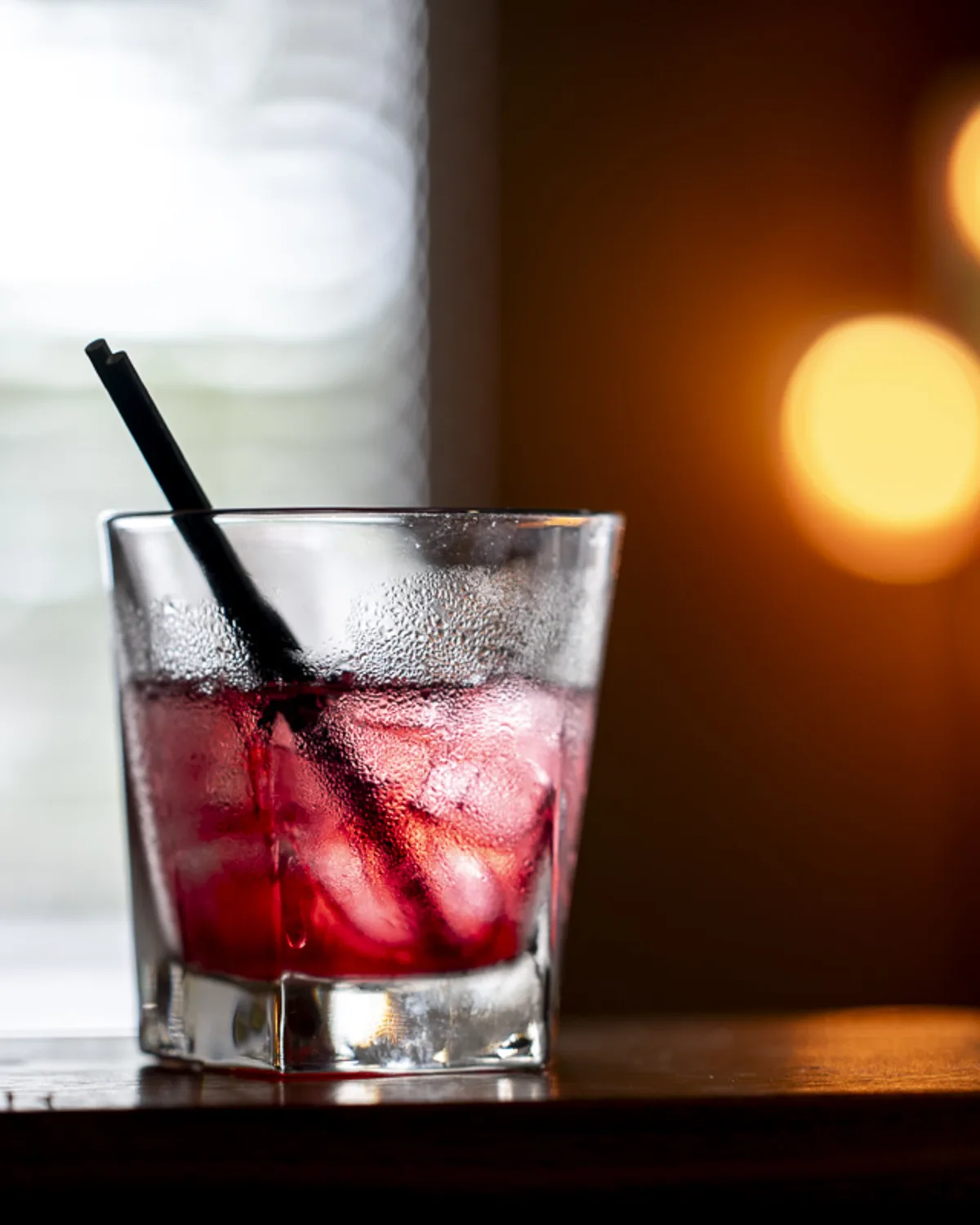 Close-up of a condensation-covered glass of red cocktail on a wooden ledge,
lit with soft window light and warm ambient bokeh during an indoor event