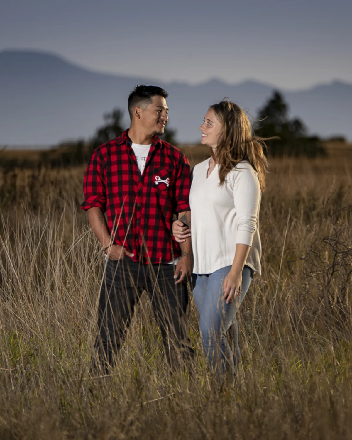 Smiling interracial couple walking arm-in-arm through tall grass at golden
hour, gazing at each other with soft light and mountain silhouettes in the background
during an outdoor portrait session