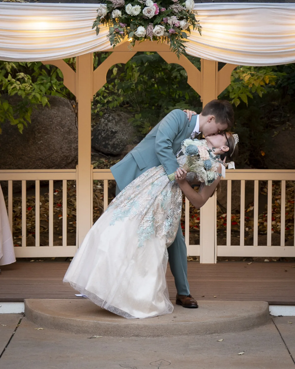 Masc-presenting lesbian in a teal suit dipping and kissing her femme bride
under a flower-adorned gazebo, with the bride’s lace dress and tiara catching soft
evening light during an outdoor wedding ceremony