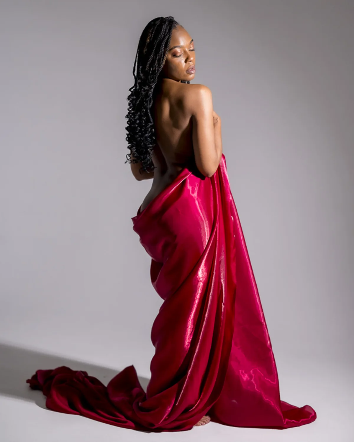 Black woman standing in profile with long braids and a red velvet drape,
softly lit in a studio boudoir session focused on elegance and self-expression