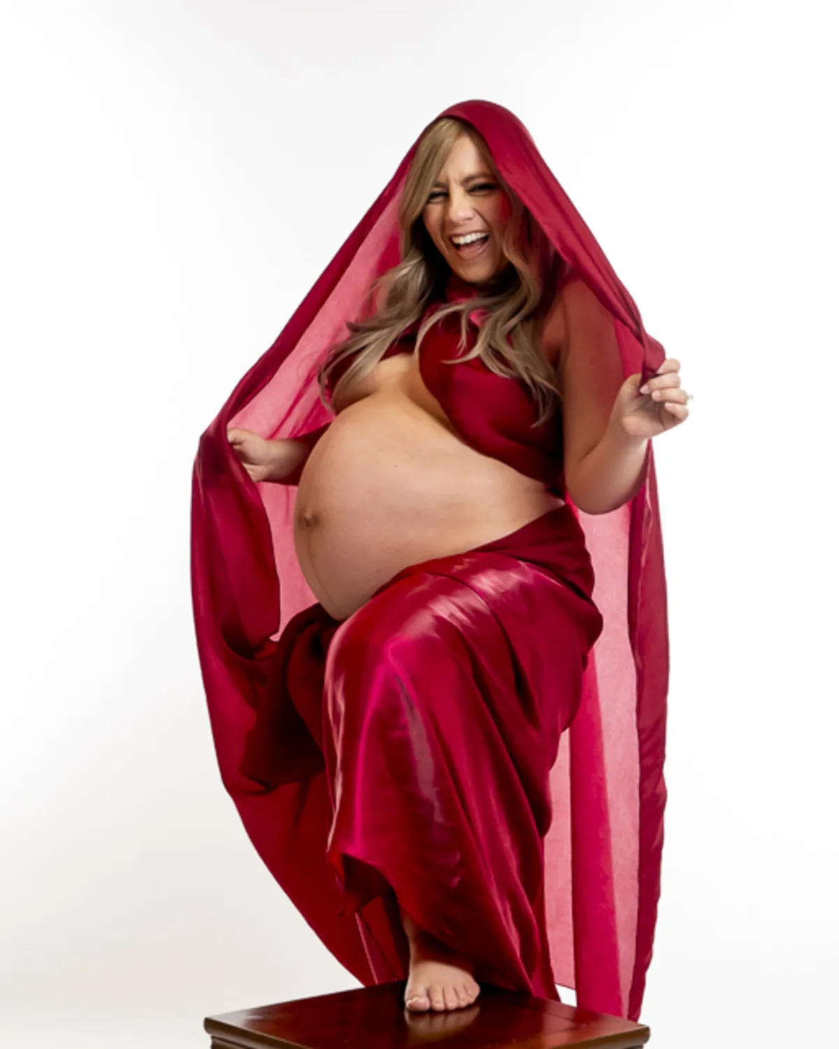 Pregnant woman laughing joyfully in a flowing two-piece gown, lifting her
hood playfully in a studio setting