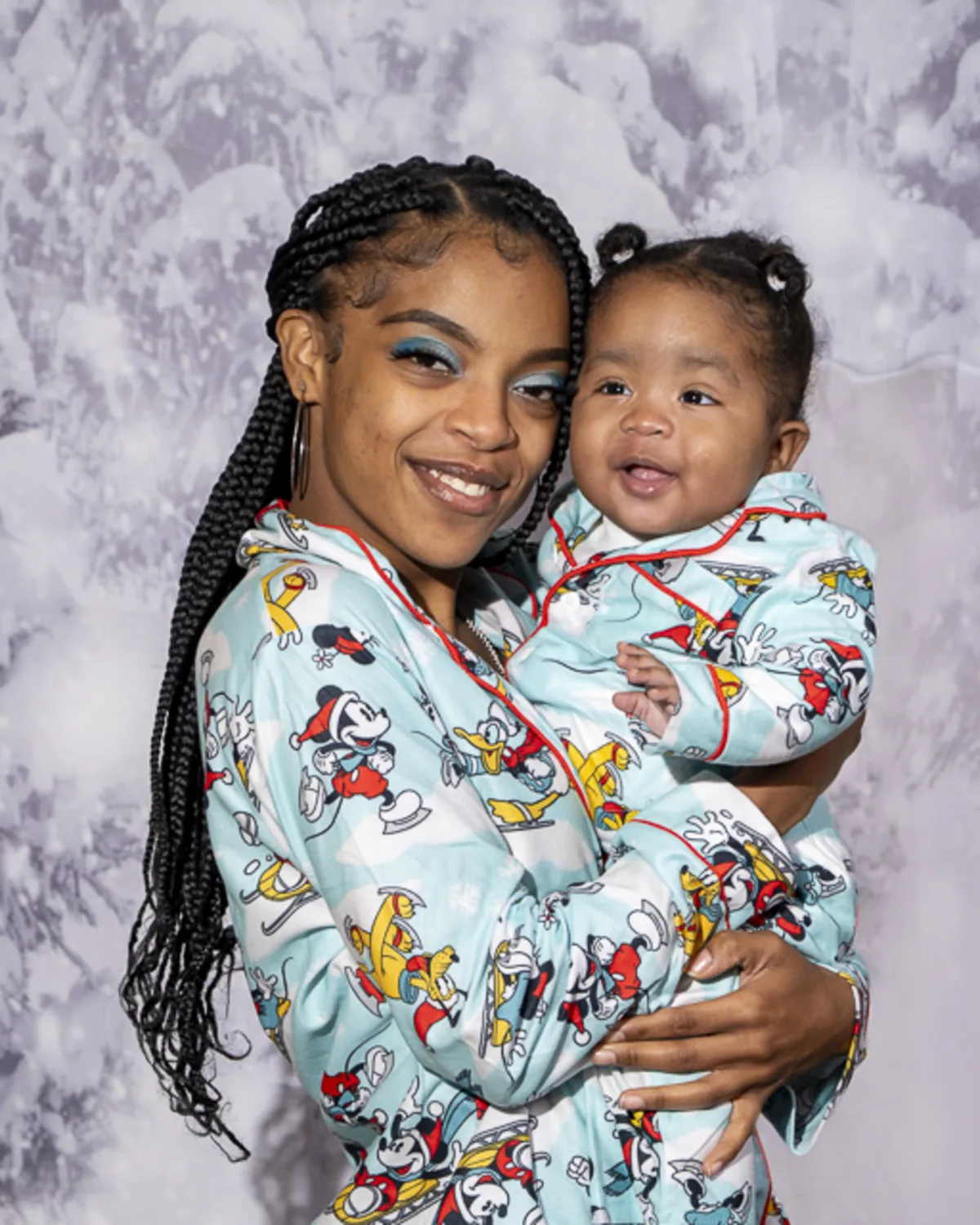 Smiling Black mother and baby wearing matching Mickey Mouse pajamas in
front of a snowy backdrop