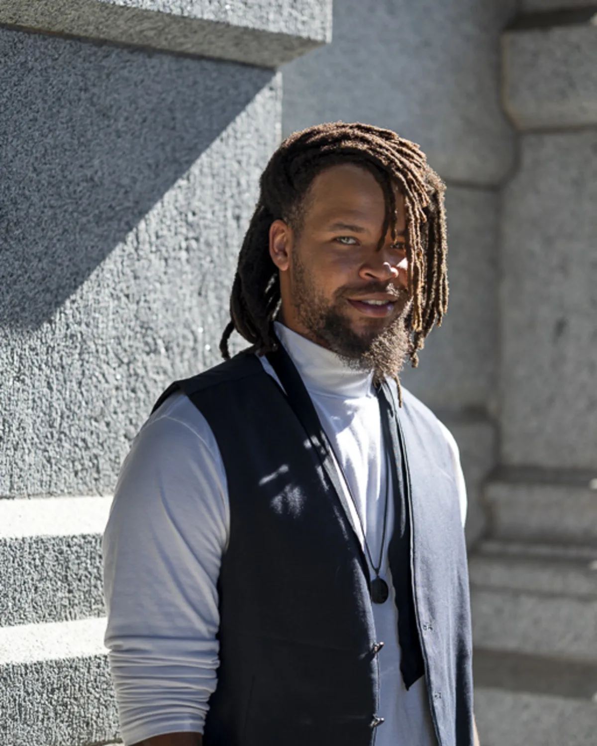 Black male model in a navy vest and turtleneck posing against a gorgeous
sunlit granite building
