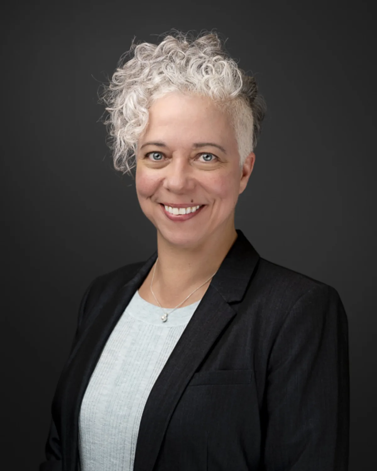 Professional headshot of a smiling white woman with short silver curls,
wearing a dark blazer against a dark gray background