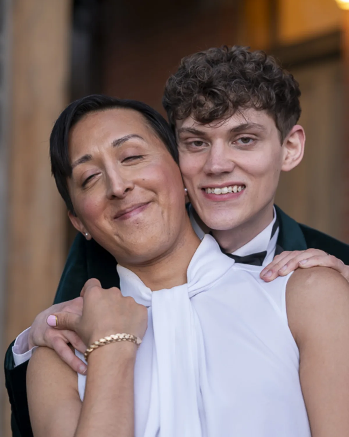 Two queer partners holding each other affectionately, dressed in
formalwear and smiling gently outdoors
