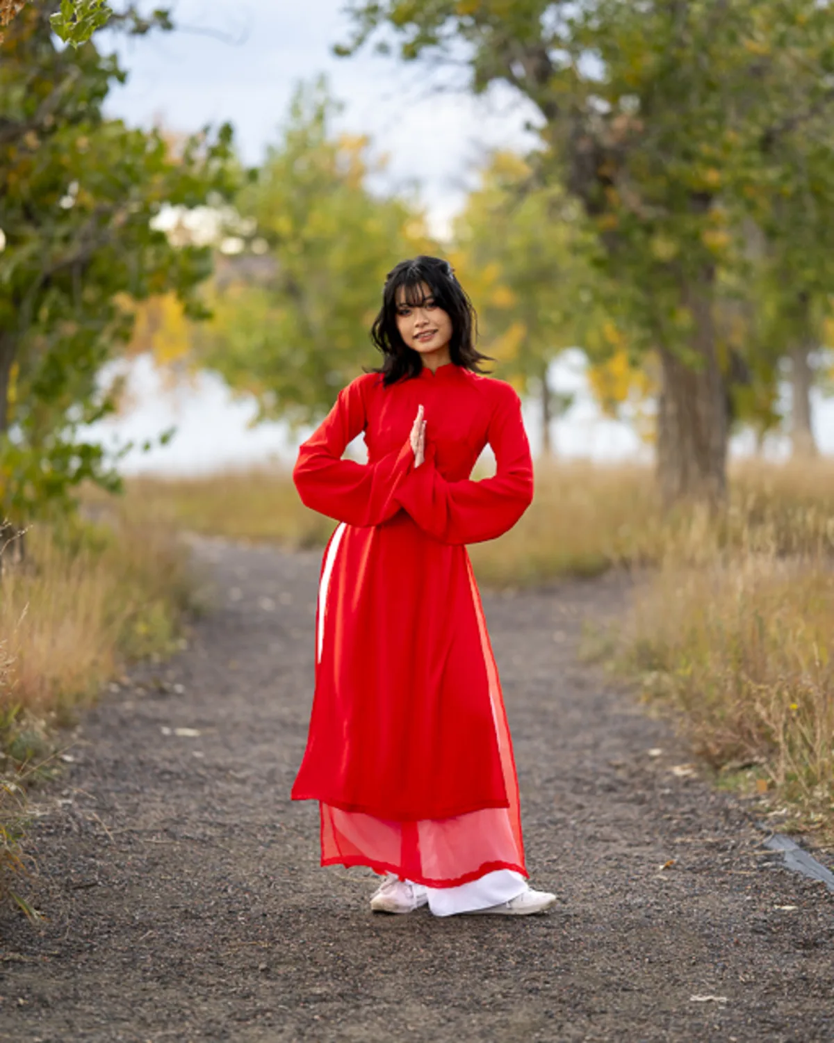 Vietnamese high school senior in a red áo dài, standing on a tree-lined
path in autumn