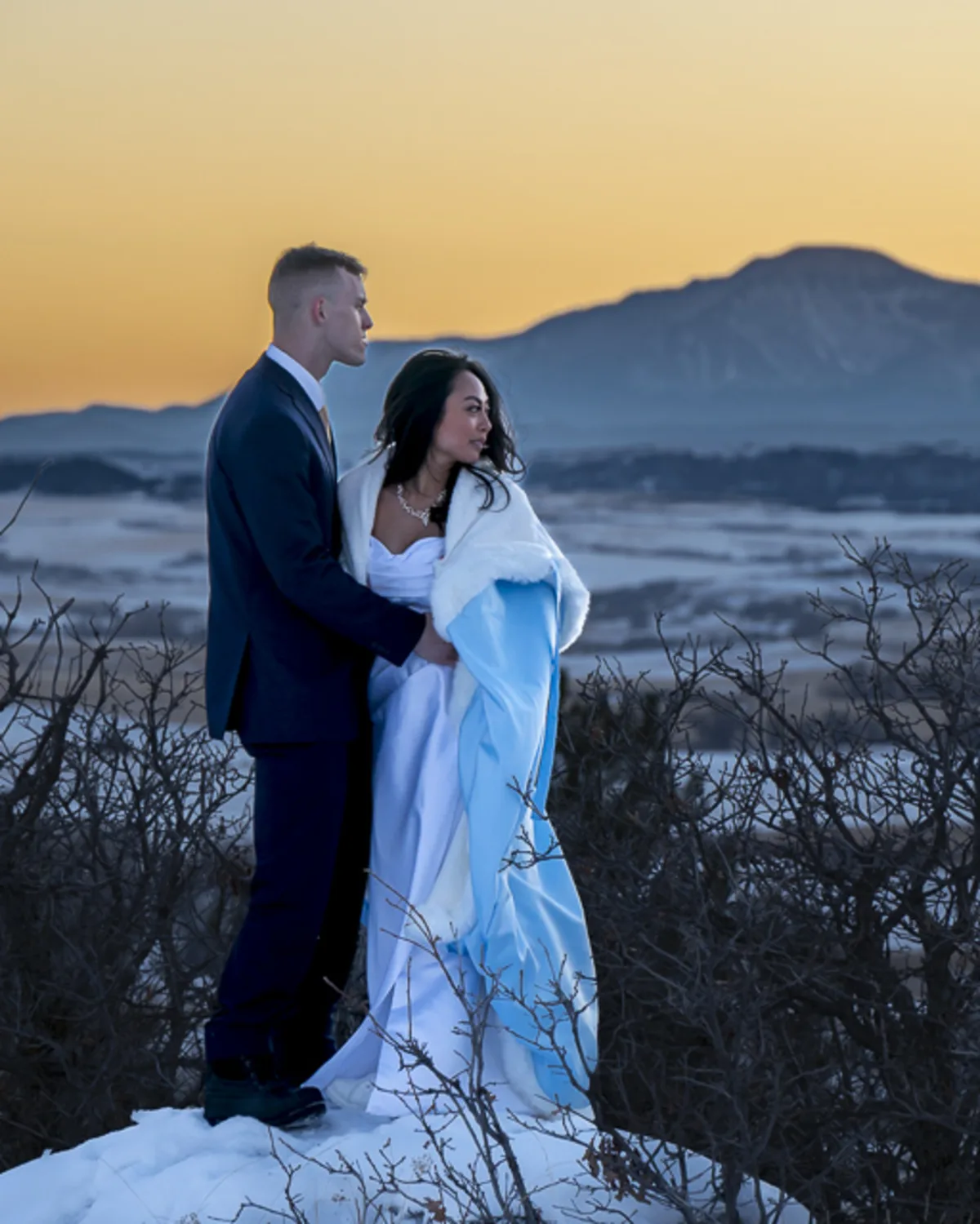 A mixed race bride and groom stand together in the snow at sunset, wrapped
in a blue and white cloak with mountains in the background
