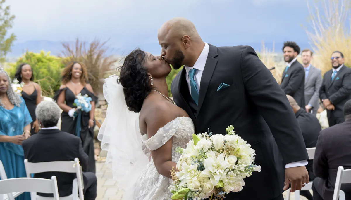 Black bride and groom share a kiss at the end of their wedding ceremony,
surrounded by joyful guests and wedding party.