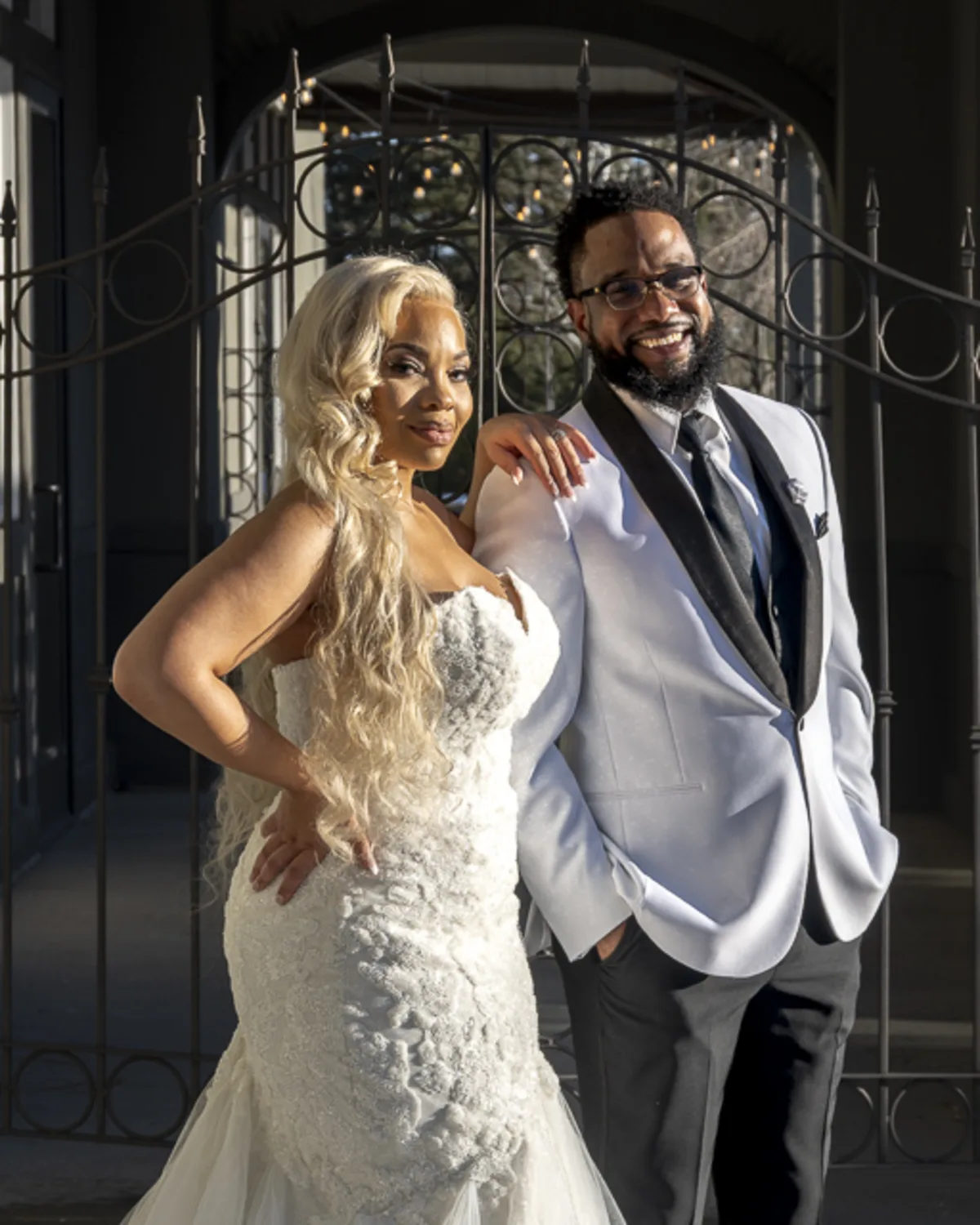 Stylish, Black, bride and groom pose confidently in formal wedding attire,
lit by golden sunlight in front of ornate ironwork