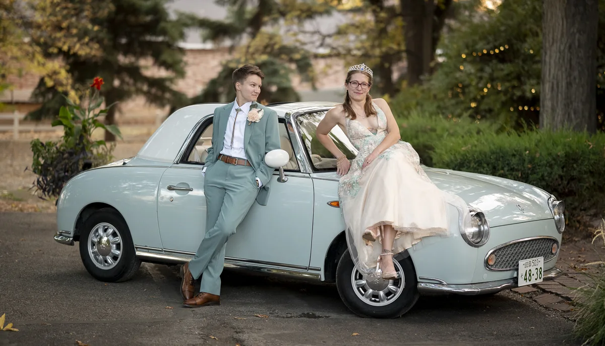 Lesbian wedding couple posing with a vintage mint green car—one wearing a teal
suit, the other in a cream and aqua gown with a tiara.