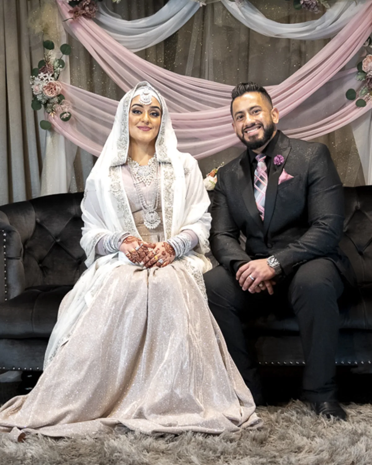 Indian bride and groom sit side by side on a velvet couch, dressed in
traditional wedding attire, with a floral and draped fabric backdrop behind them