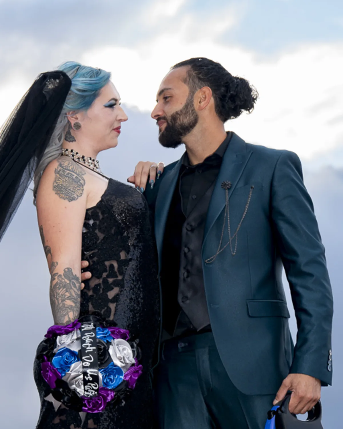 Tattooed bride in a black dress and groom in a dark teal suit share a
loving gaze under dramatic skies, holding a gothic-style bouquet.